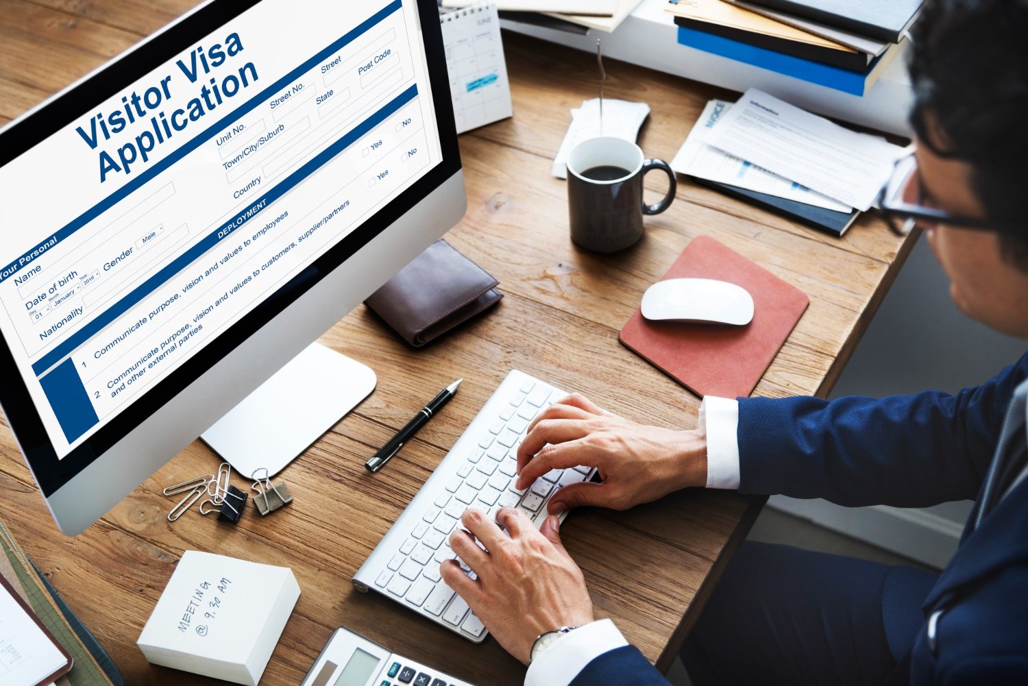 Person working at a desk on a visitor visa application form on a desktop computer, with office supplies nearby, including a cup of coffee, notepad, pen, and binders.