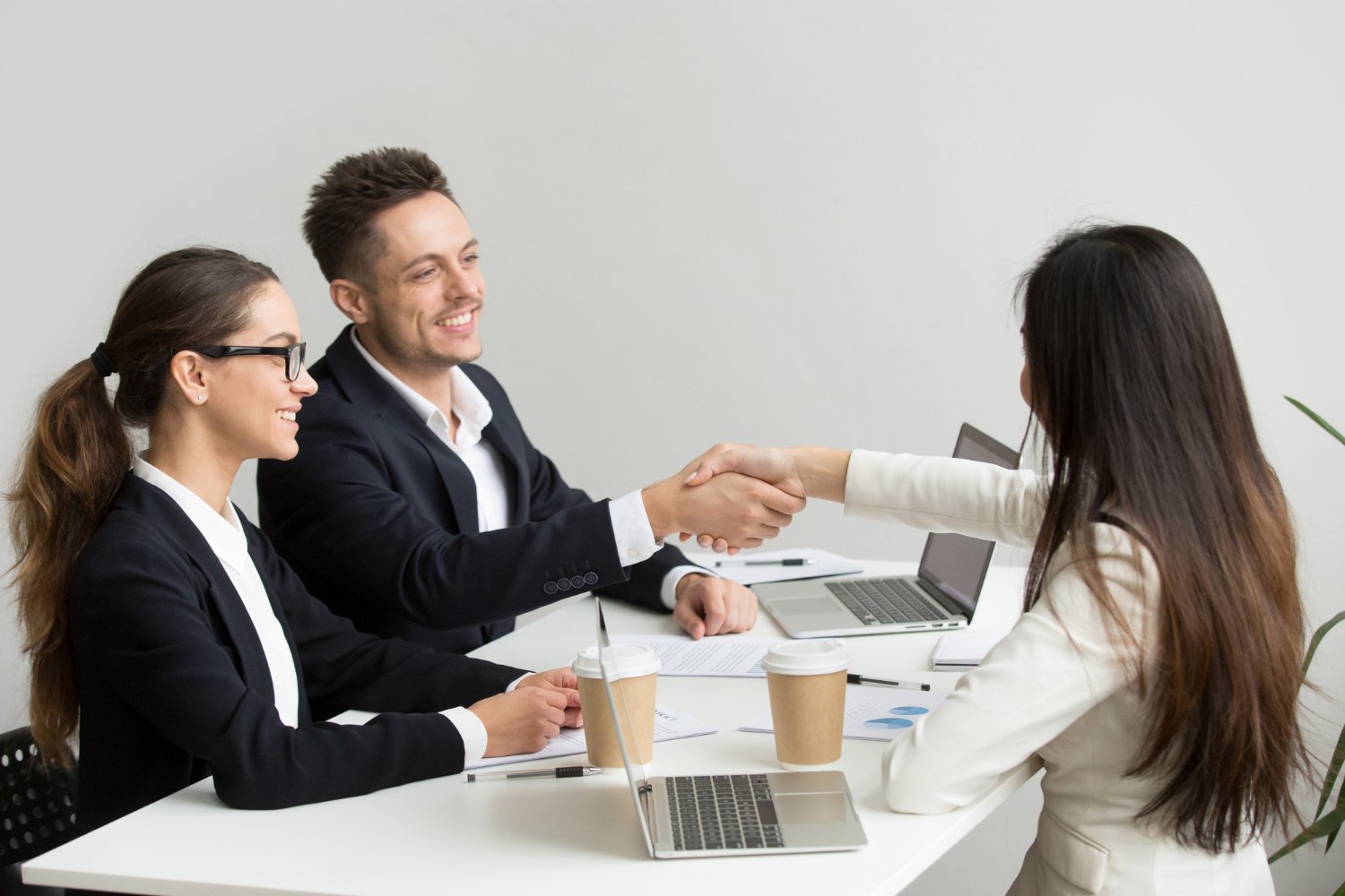 Two business professionals, a man and a woman, shaking hands with another woman across a table during a meeting, with laptops and coffee cups present.