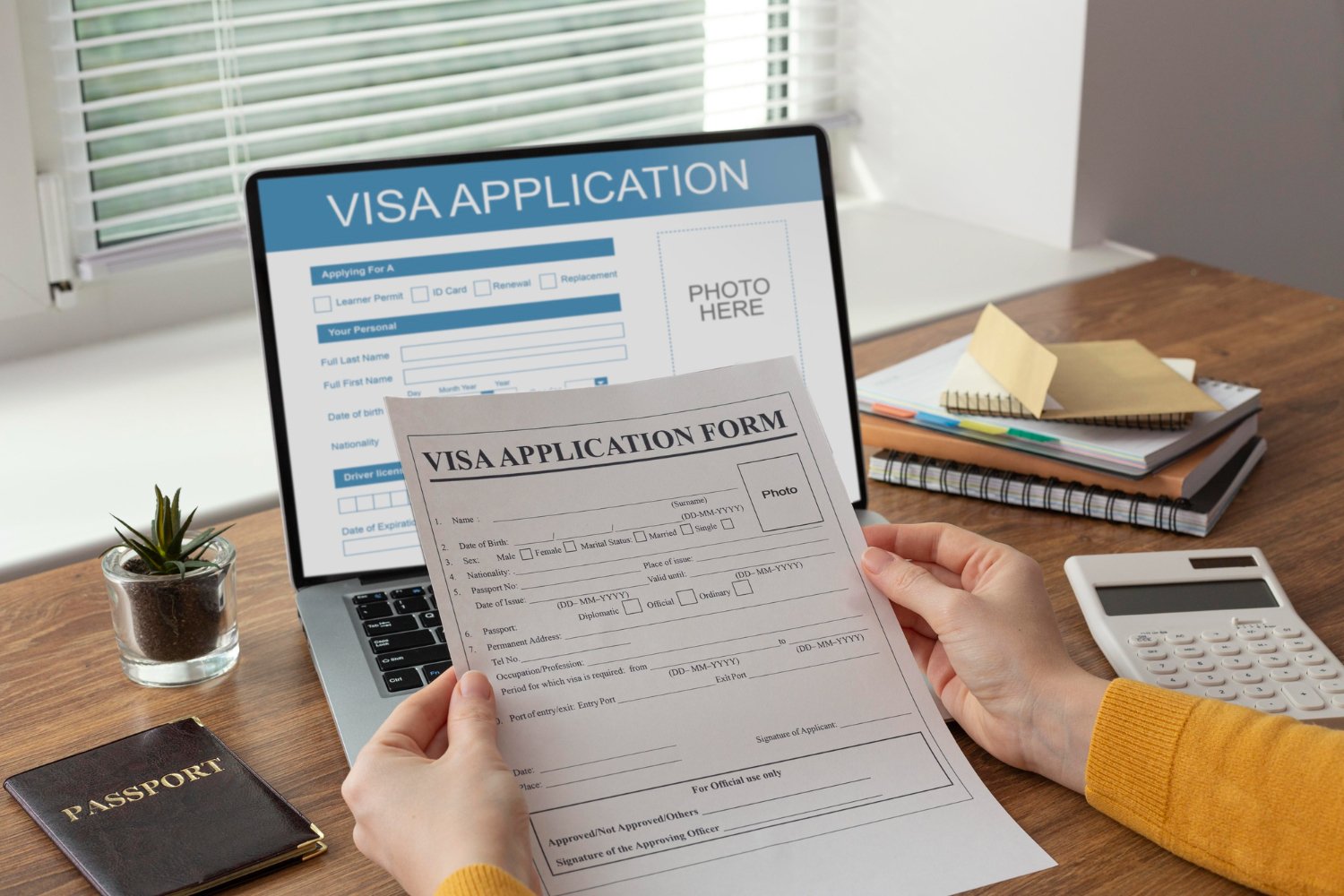 Person reviewing a visa application form at a desk with a laptop displaying an online visa application, a passport, a calculator, a small potted plant, and a stack of notebooks and papers.